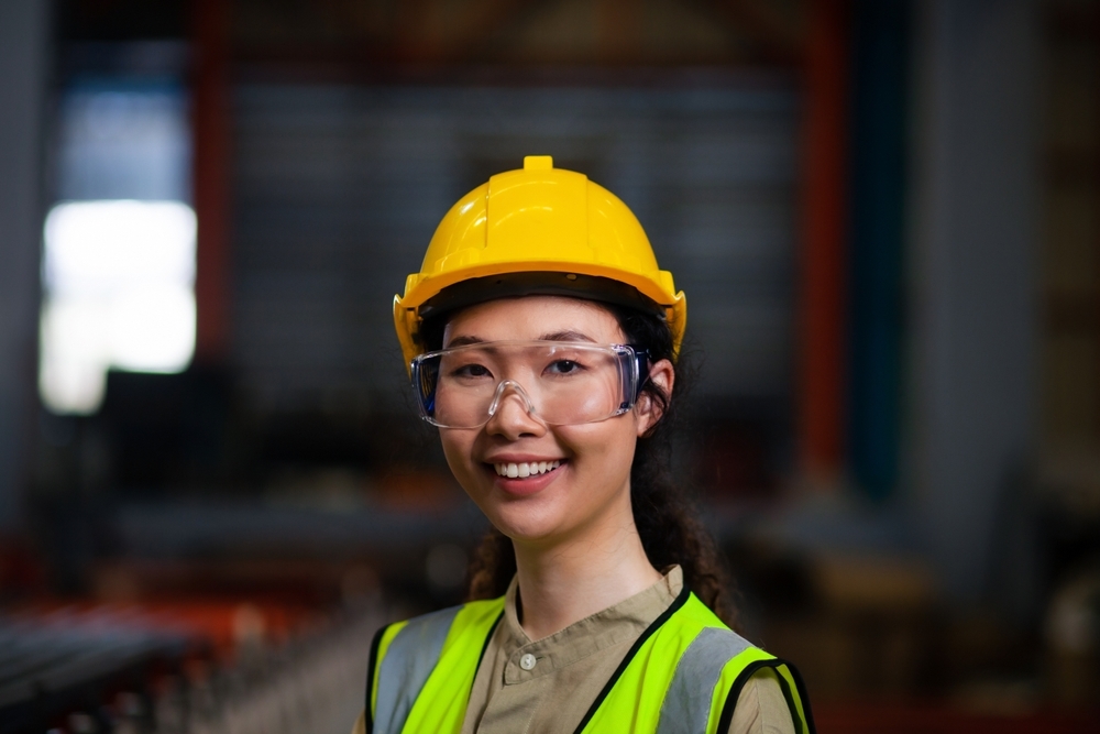 Portrait of a confident female worker in a safety vest with a hard hat smiling at the camera in a sheet metal factory.