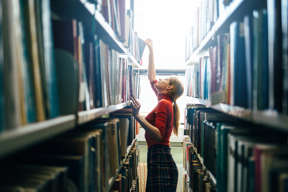 Young female librarian searching books and taking one book from library bookshelf.