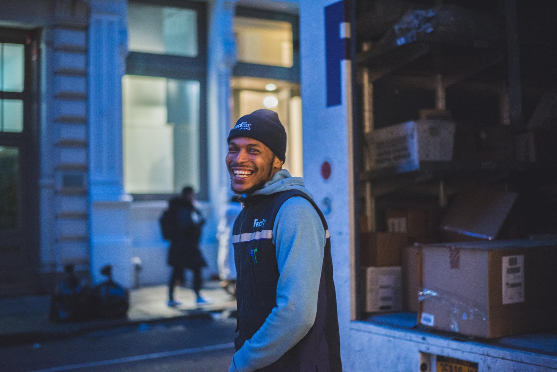 A delivery driver wearing a beanie smiles while unloading packages from a truck.