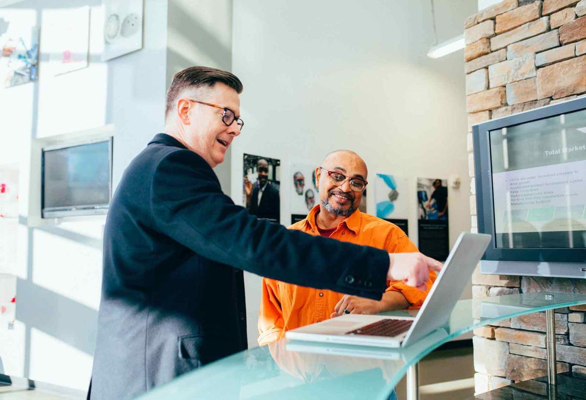 Two men standing in front of a desk, one pointing to a laptop while giving the other man feedback at work