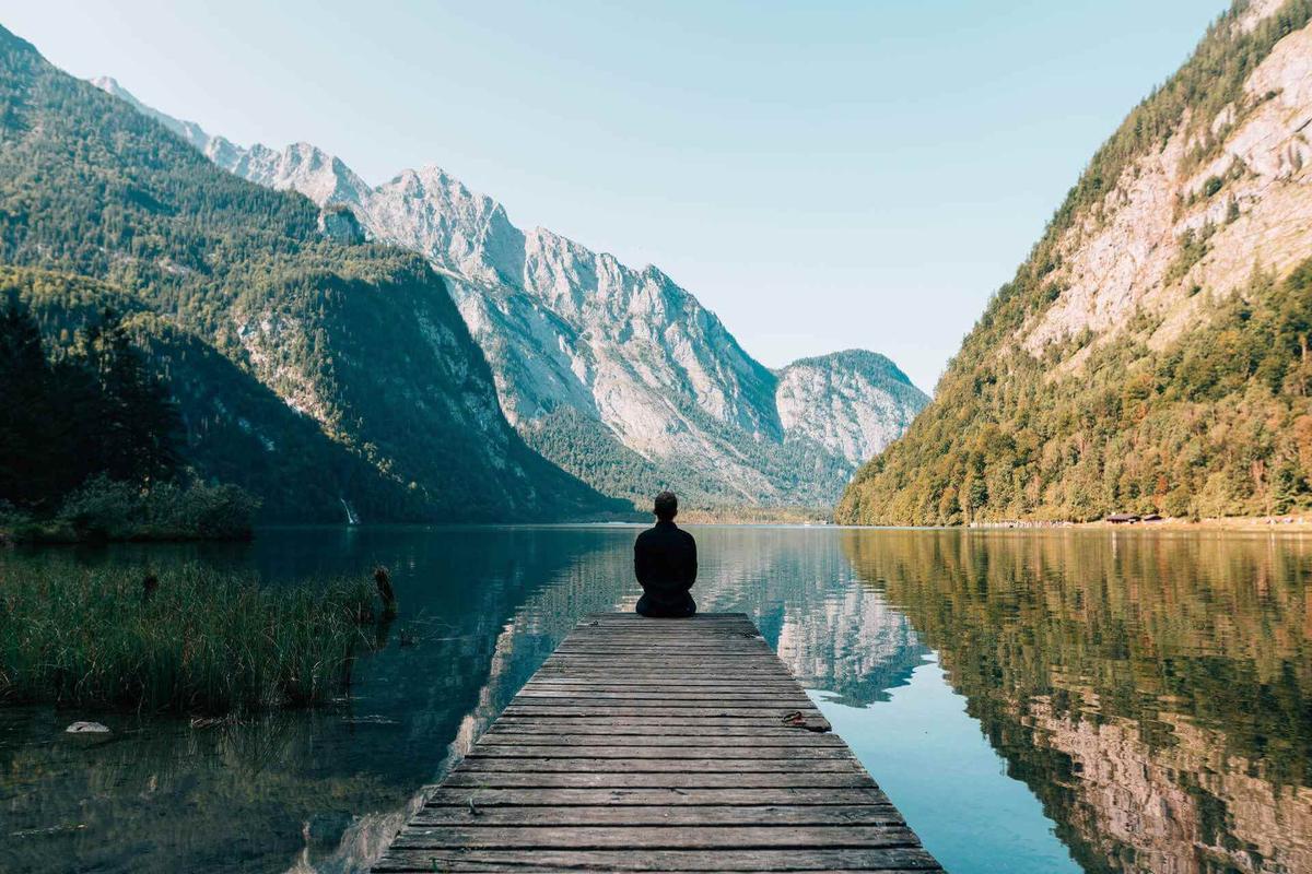 Person meditating on gangplank surrounded by lake and mountains