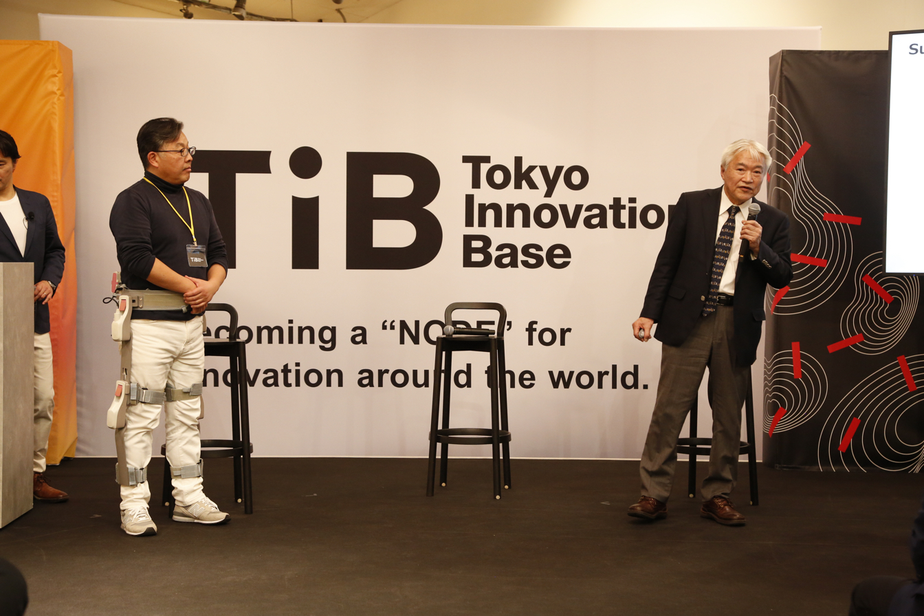 Mainichi Future Creation Lab's CEO Tamotsu Takatsuka (left) tries on the robotic suit designed to make it easier for elderly people to walk as its developer Assist Motion's CEO Minoru Hashimoto explains the product for the audience.&nbsp; &nbsp;