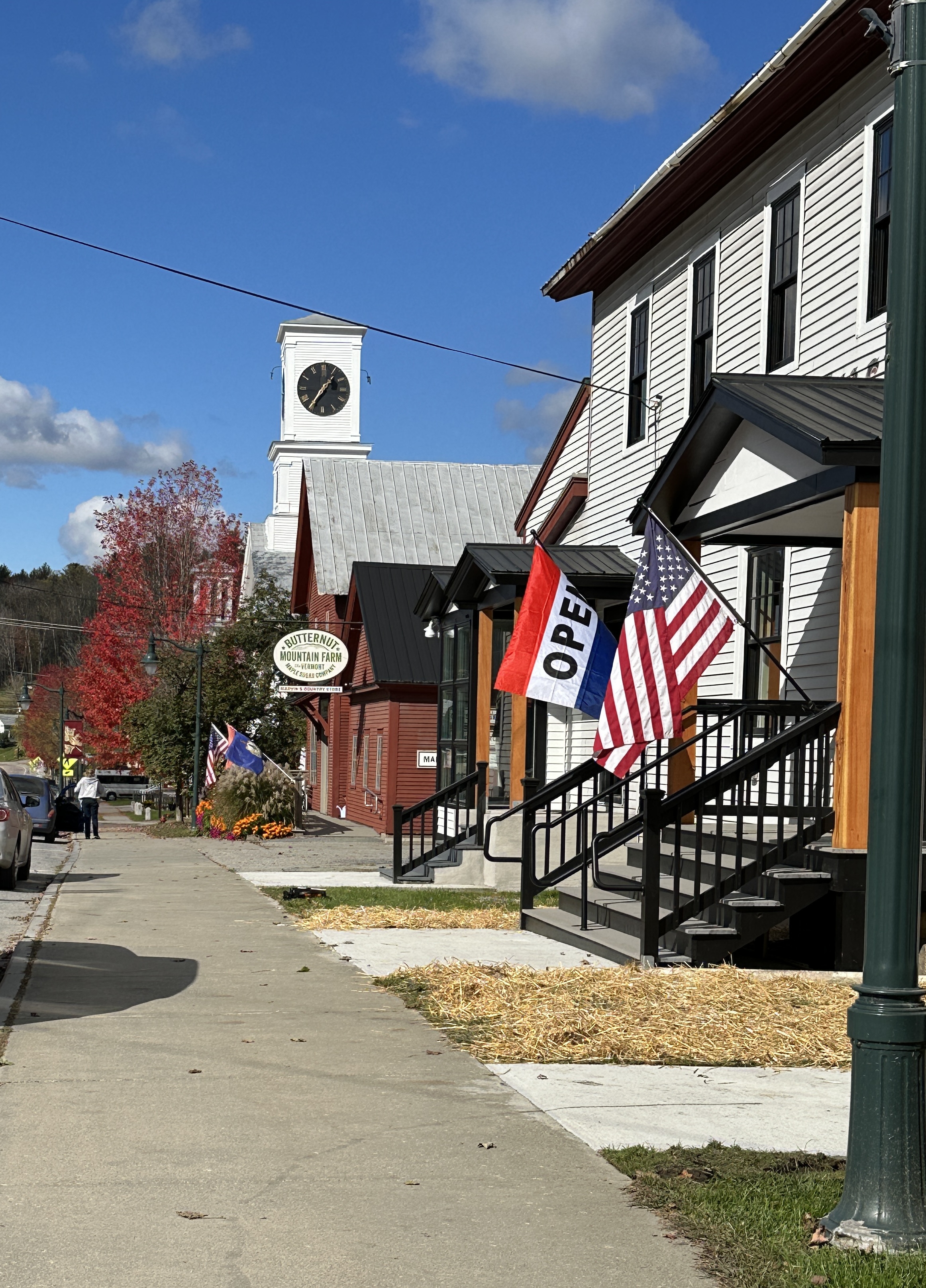 Main Street in Johnson on an autumn afternoon. The clock tower rises above the rooflines, an OPEN flag and an American flag hang outside the storefronts, and a red maple is in full color.