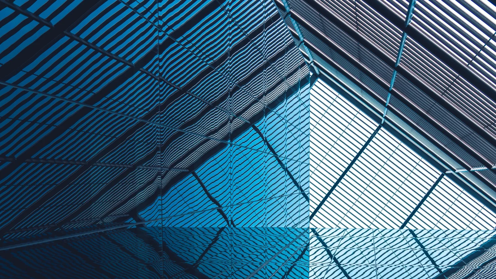 Abstract, angular architecture. Diagonal, blue, metallic beams viewed from inside an office building looking up. The image is partially mirrored.