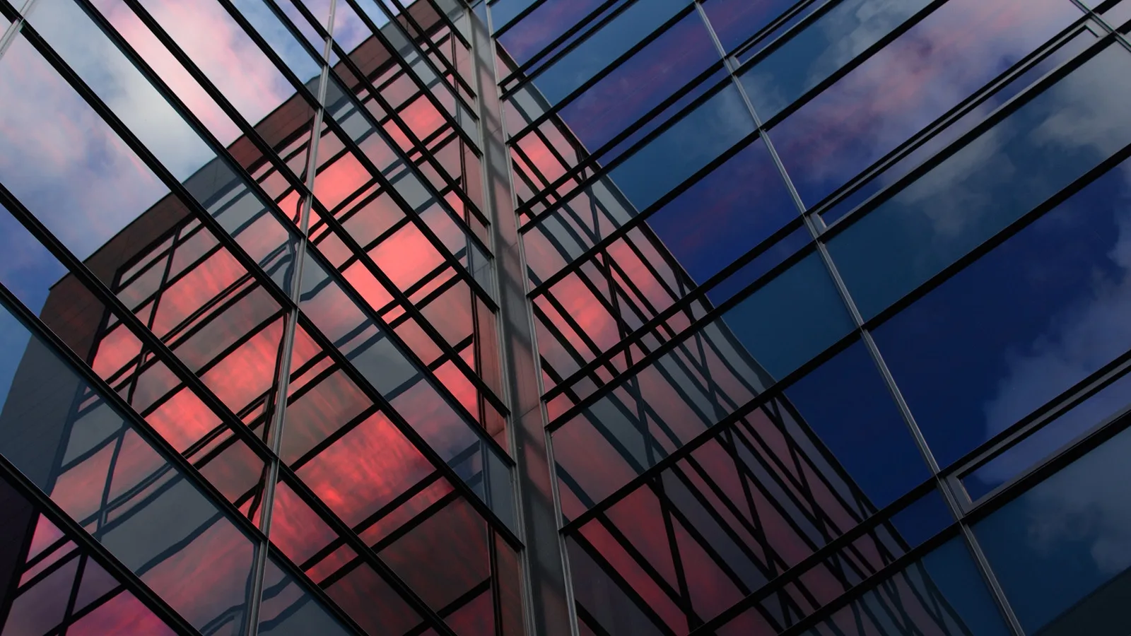 The mirrored exterior of an office building with the sunset captured by the overlapping glass panels. Colours: pink clouds and blue sky are reflected.