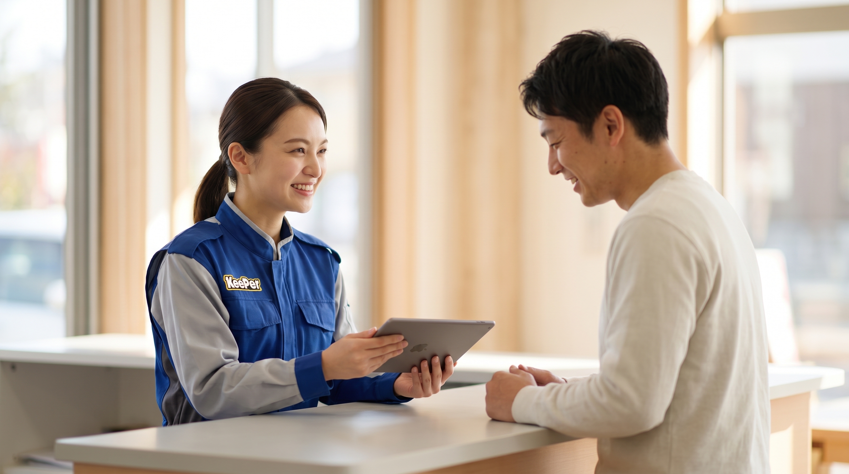 タブレットを見ながら笑顔で相談するスタッフと車のオーナーの様子。背景をぼかし、柔らかな自然光と親しみやすい暖色系で、和やかに会話する2人を横から捉えています。