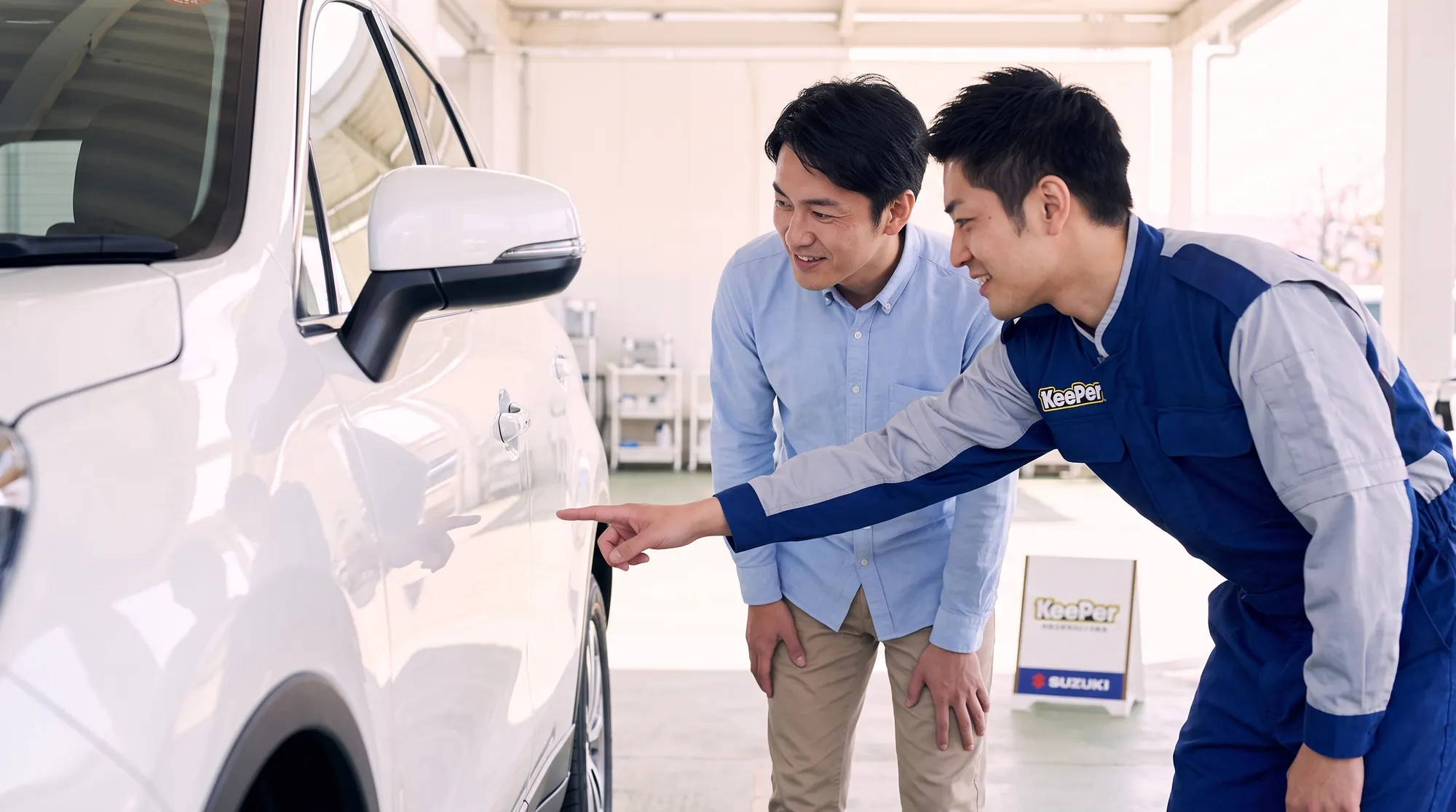 白い車のドア付近を指差して、塗装の状態を一緒に確認し合うオーナーとスタッフの様子。柔らかい自然光に包まれた、和やかで親しみやすい雰囲気のミディアムショット。