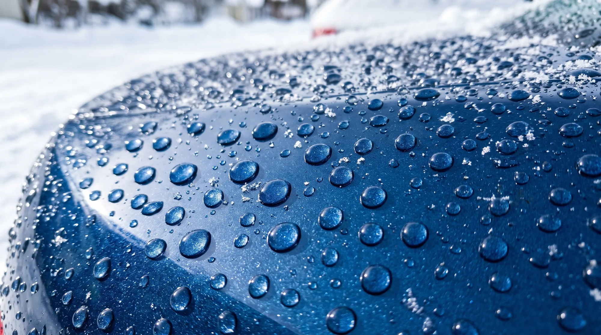 深いブルーの車のボディパネル上で、雪や氷がコーティング効果により美しい水滴となって弾かれているクローズアップ。水滴の透明感と塗装の質感が際立っています。