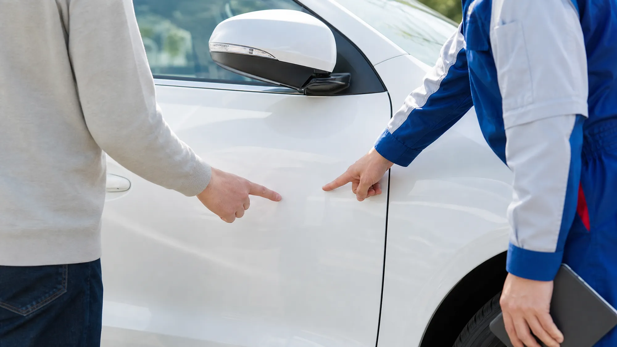 白い車のドアパネル付近で、青い制服のスタッフと顧客が塗装面を指差して状態を確認する手元のクローズアップ。明るい日差しの下での、爽やかで清潔感のある様子です。