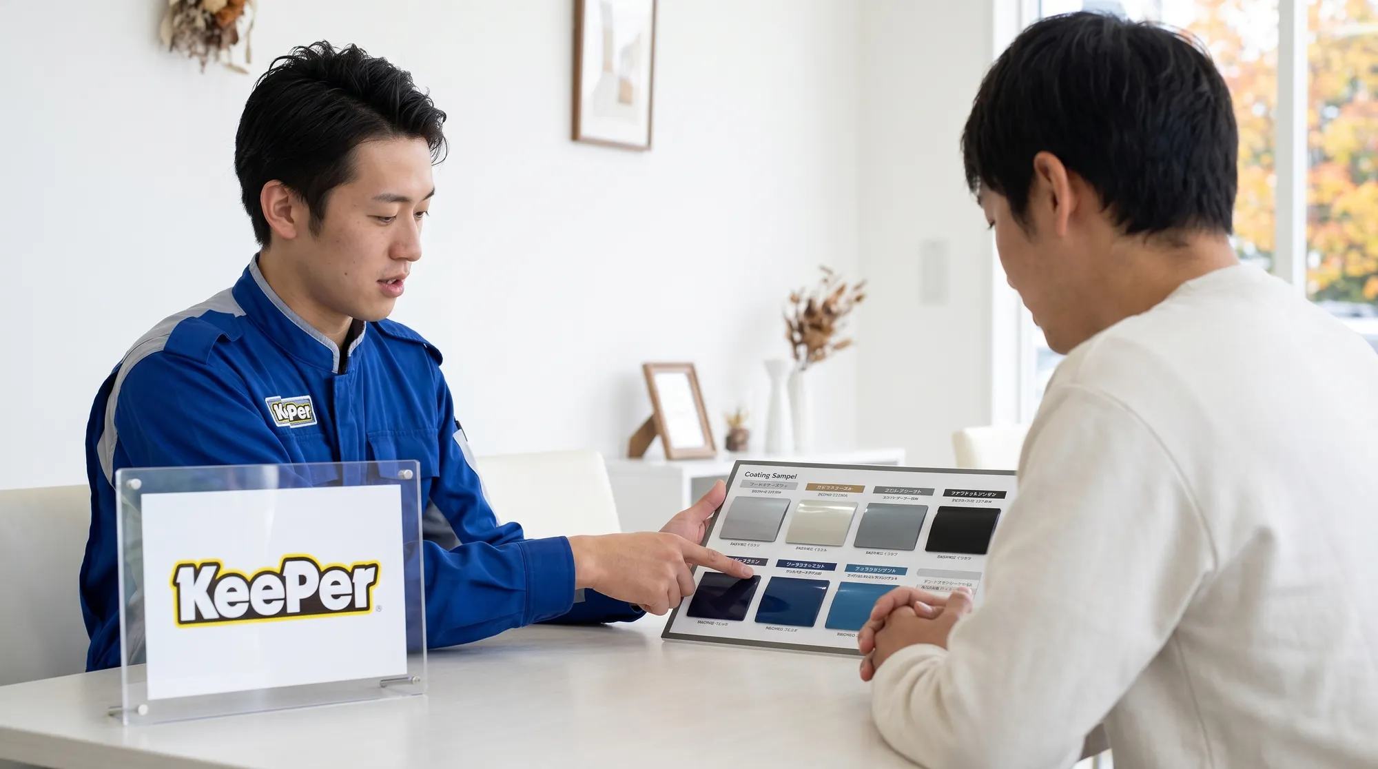清潔な白い壁を背景に、青い制服のスタッフと顧客がテーブル越しに打ち合わせをしている様子。スタッフがパネルを指差して説明する、信頼感と落ち着きのあるシーンです。