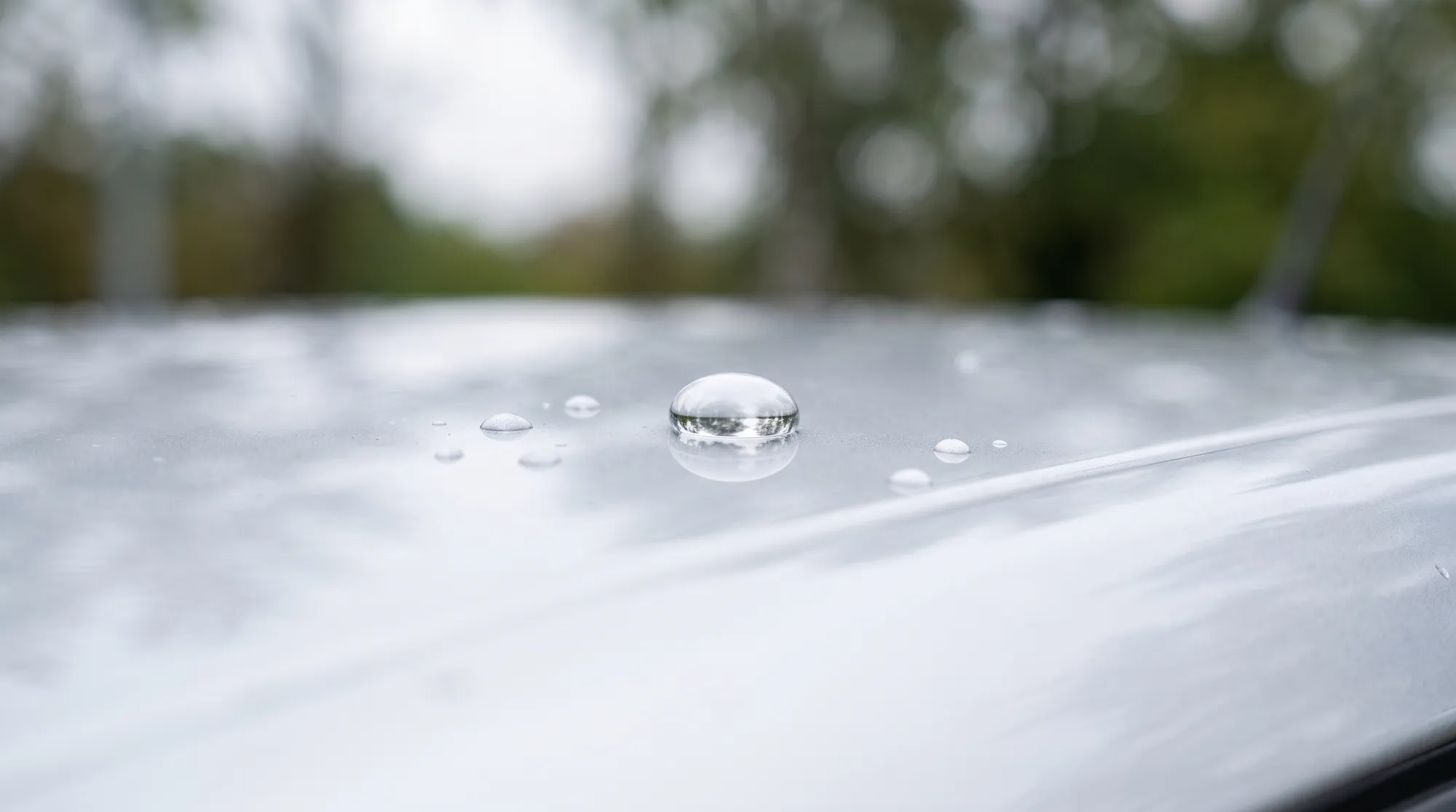 雨上がりの屋外で、車のルーフに乗った美しい水滴のクローズアップ。コーティングの強力な撥水効果で、水滴が真ん丸な球体となってボディを滑り落ちようとしているシーン。