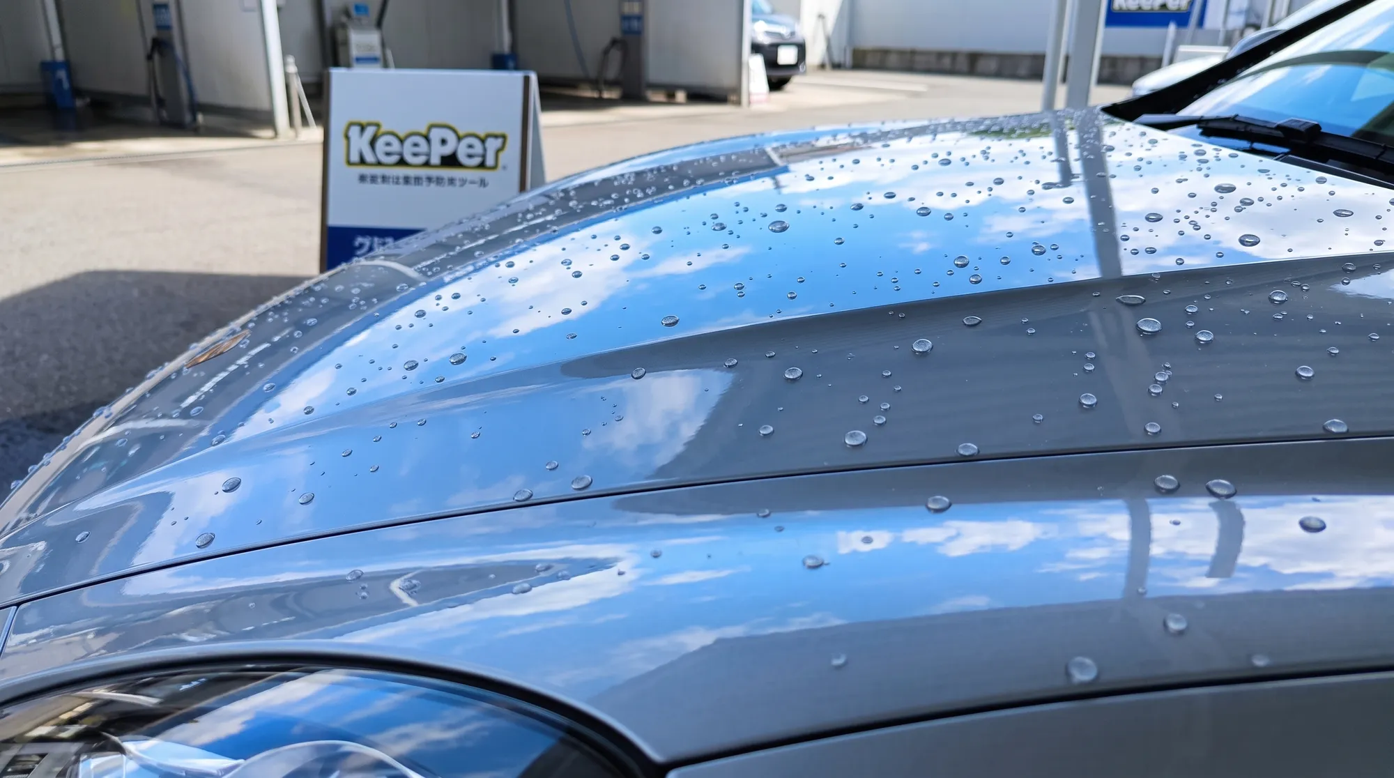 爽やかな自然光の下、コーティングされた車のボンネットで美しい球状の水滴が弾いている様子。鏡のようなボディに、鮮やかな青空と白い雲が反射しているクローズアップ。