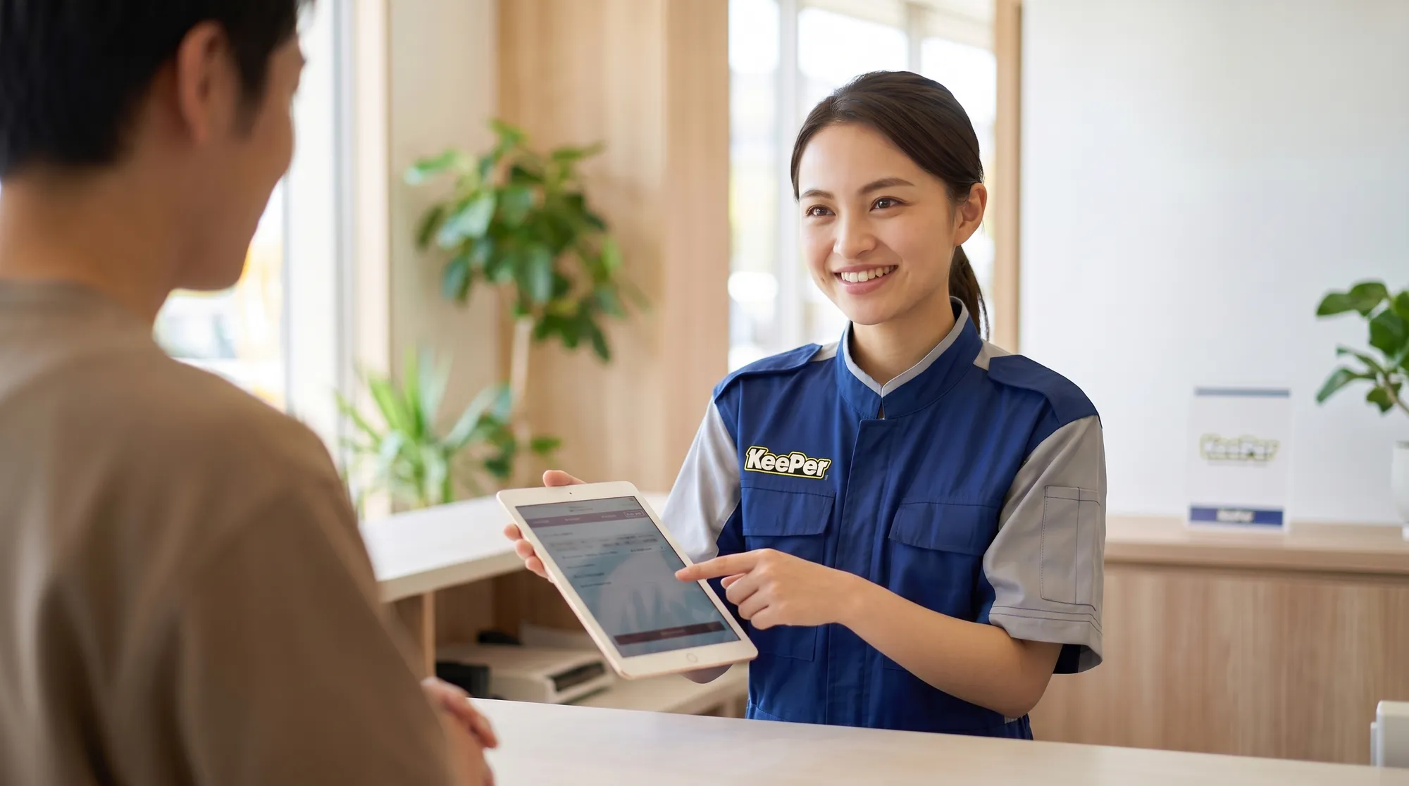 明るい店舗の受付で、スタッフがタブレット画面を見せながら顧客に案内している様子。笑顔で説明するスタッフを顧客の肩越しに捉えた温かみのあるクローズアップ。