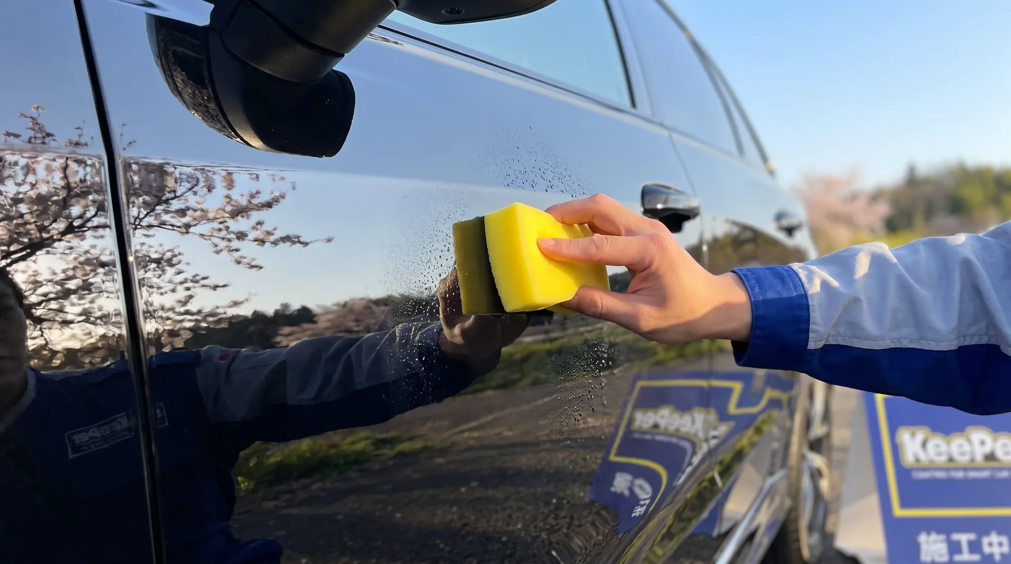 汚れを除去して美しい艶を取り戻した黒い車のドアパネルのクローズアップ。スタッフが専用スポンジでコーティング剤を丁寧に塗り広げ、鏡のような深い光沢を放つ様子。