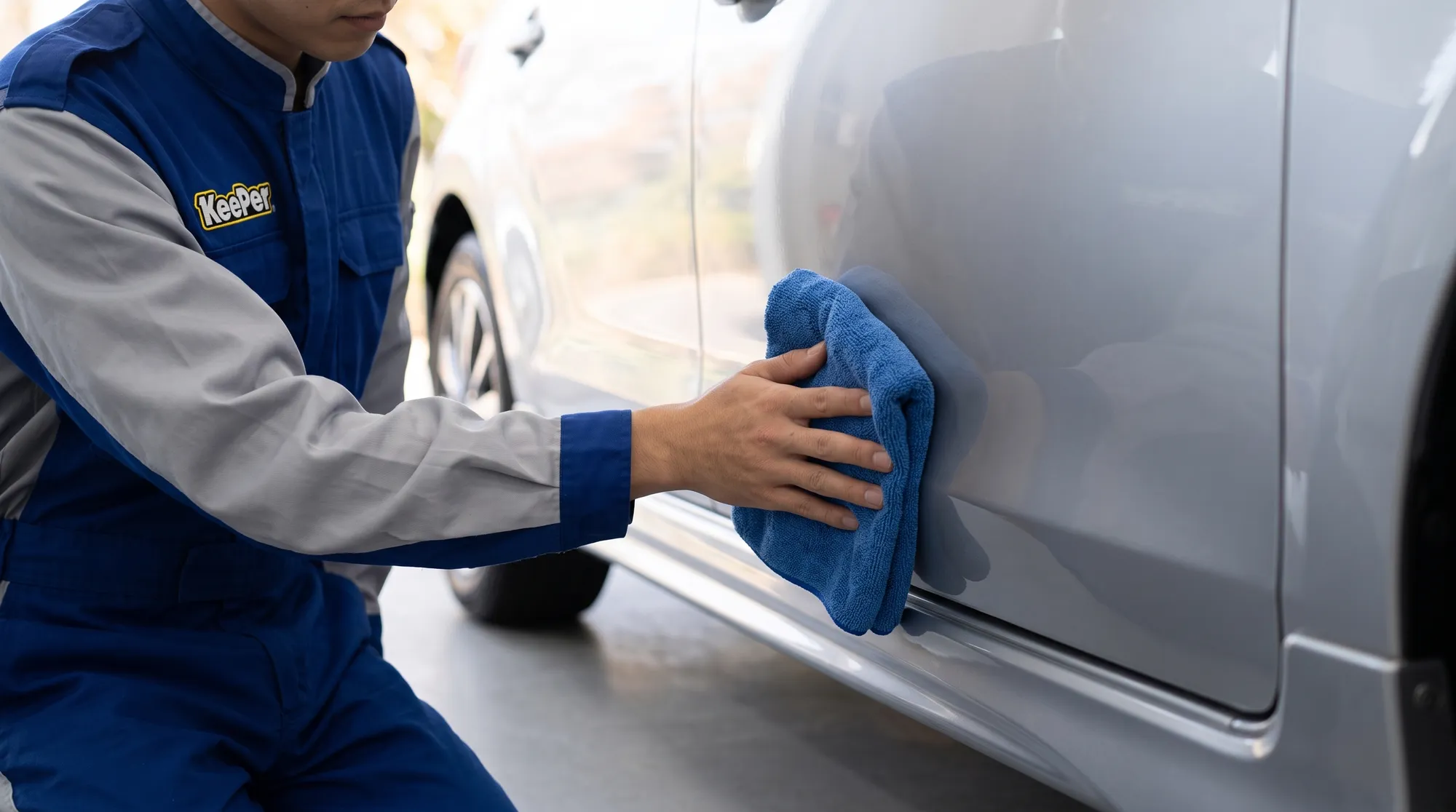 清潔な洗車ブース内で、ユニフォーム姿のスタッフが青いマイクロファイバークロスを持ち、シルバーの車のドアパネル下部を優しく拭き上げている手元のクローズアップ。