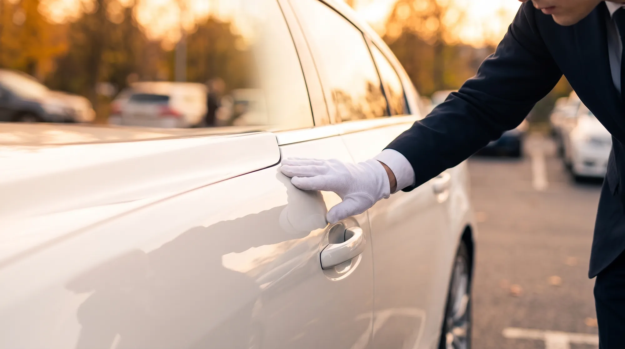 白い手袋をしたスーツ姿の人物が、白い車のサイドドアの艶を丁寧に確認している様子。夕暮れ前の暖かみのある自然光が、車の美しい曲線と深い光沢を引き立てています。
