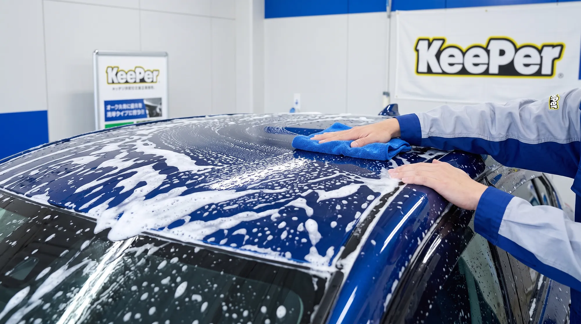 深い青色の車のルーフを、豊かな白い泡と青いマイクロファイバークロスで優しく洗車している様子。午後の柔らかい自然光が、水滴と泡をキラキラと輝かせています。