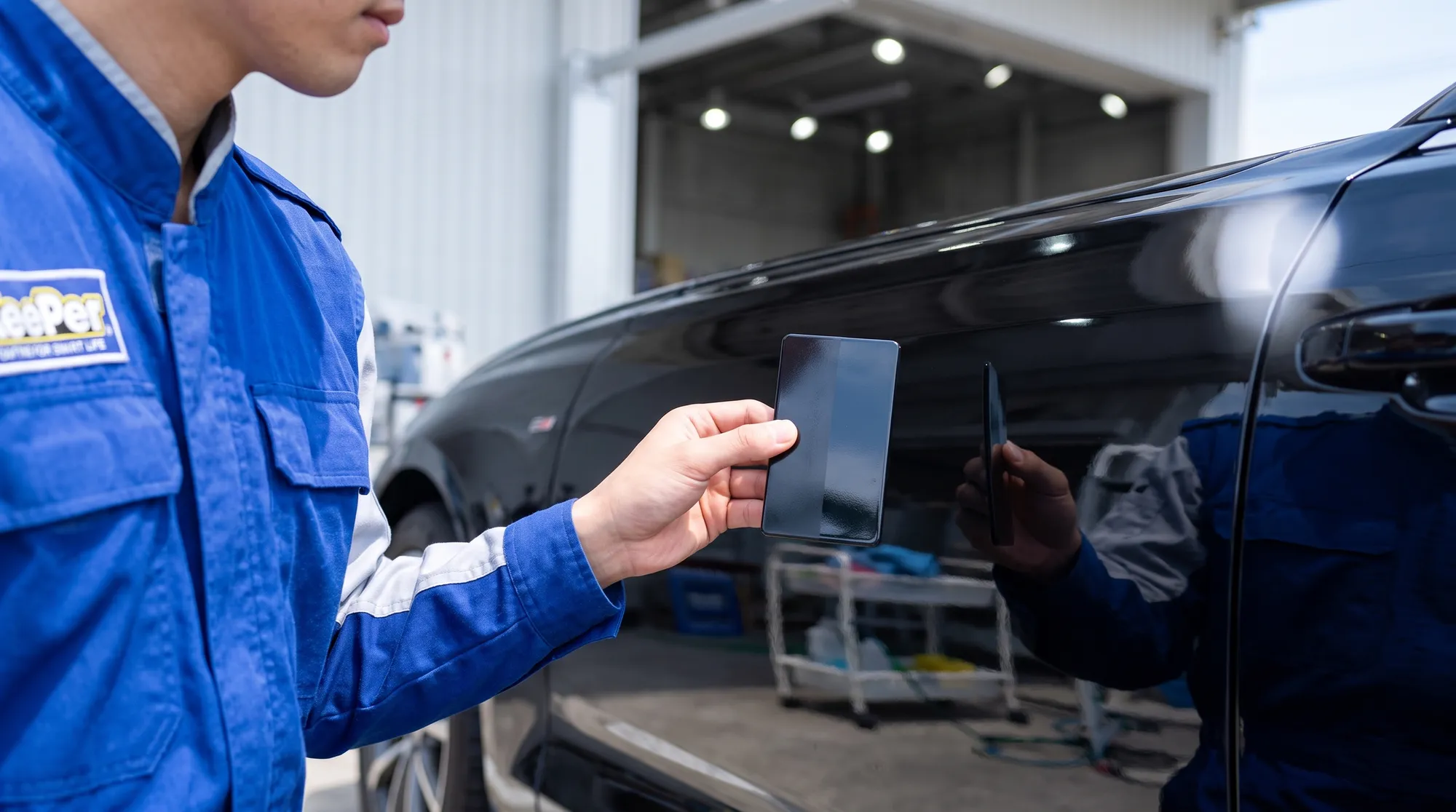 深い黒色の艶やかな車の横で、青い制服のスタッフがコーティング比較用サンプルを手に持つ様子。光がボディの美しい艶を際立たせるプロフェッショナルなクローズアップです。