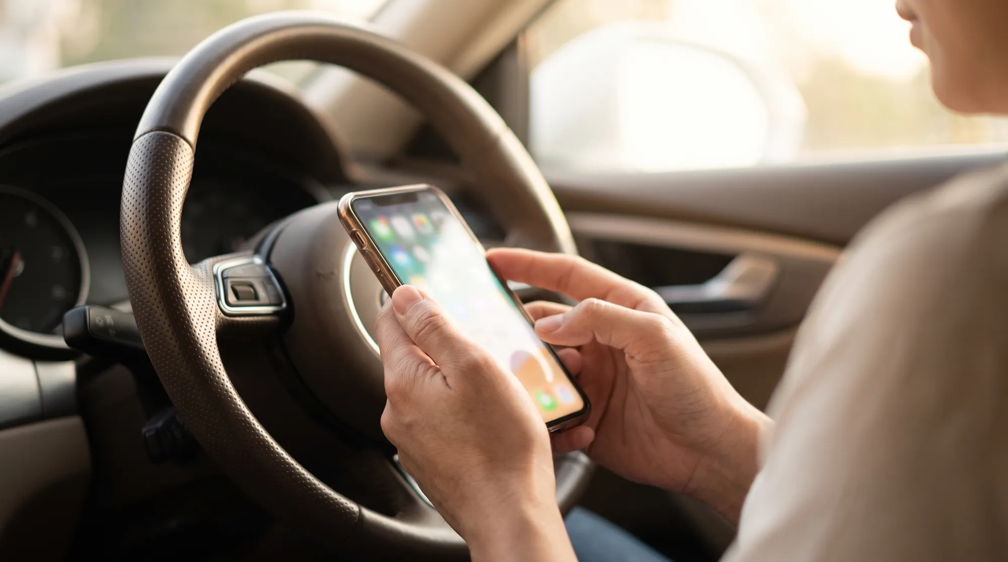 柔らかい自然光が差し込む車内で、レザーステアリングを背景にスマートフォンを操作する人物の手元のクローズアップ。温かみがありリラックスした雰囲気のシーンです。
