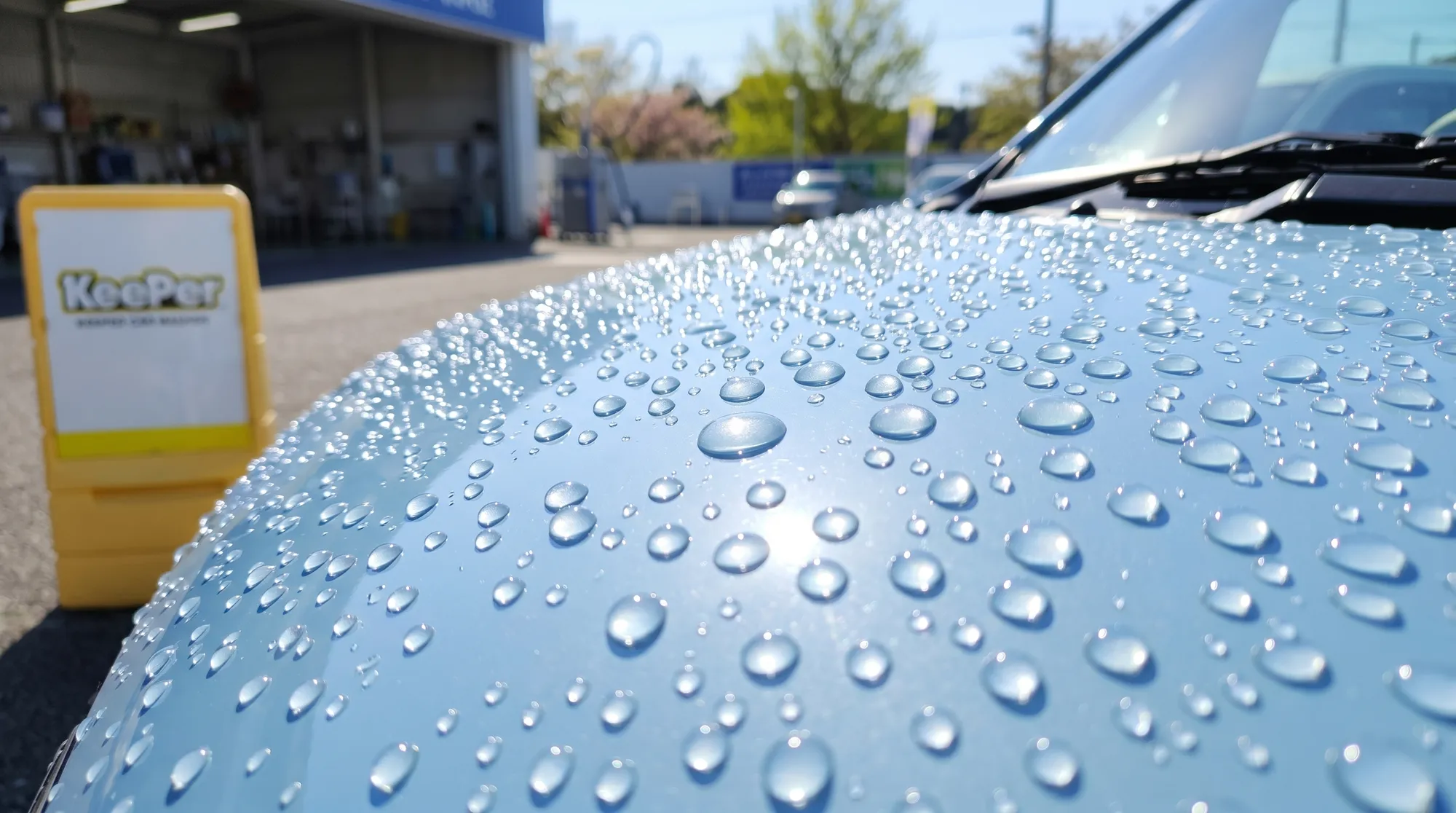 雨上がりの明るい屋外スペースに停められた、パステルカラーのコンパクトカーのボンネットのクローズアップ。表面にできた美しい球状の水玉に太陽光が反射して輝く様子。