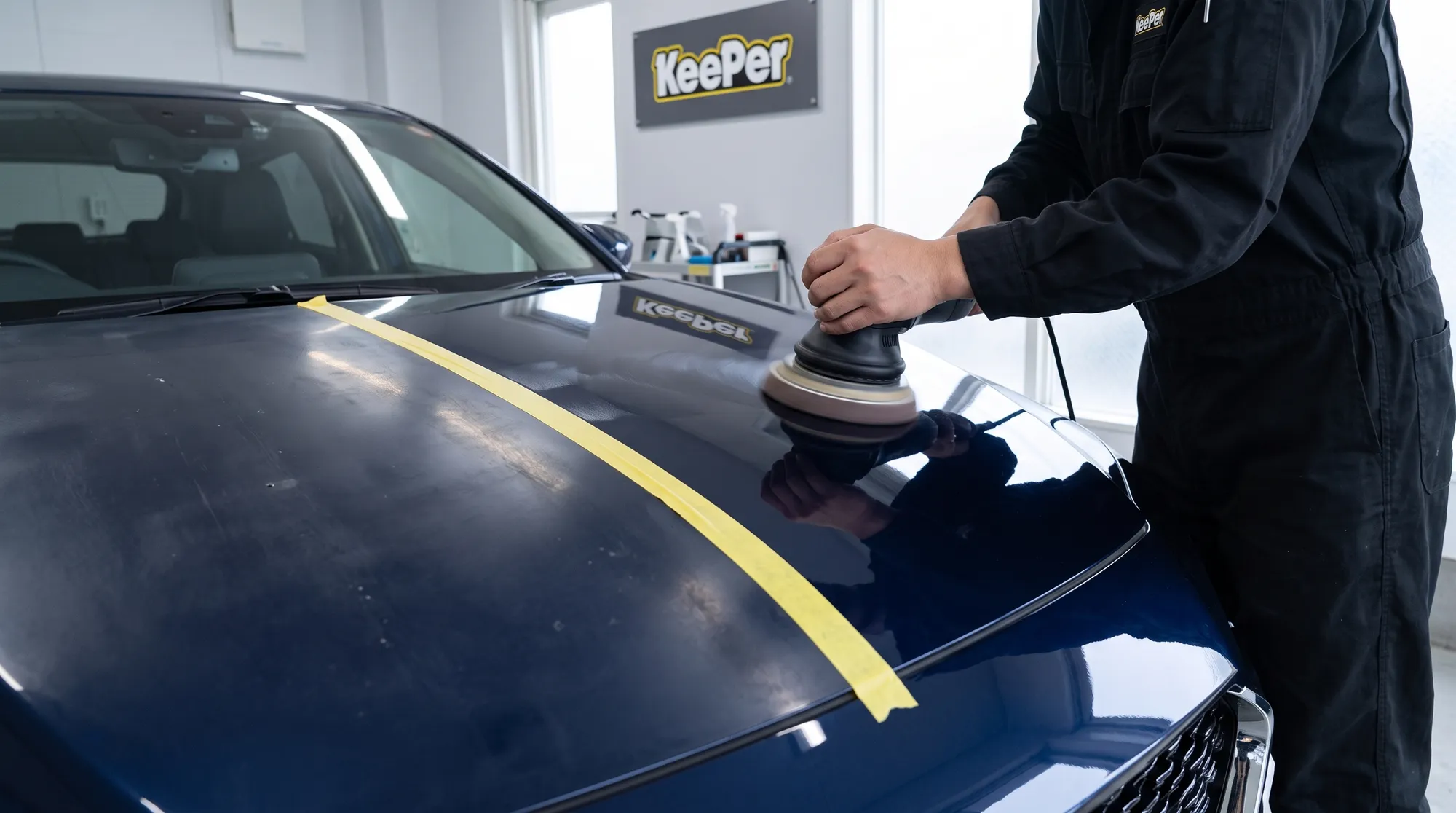 紺色の車のボンネットをポリッシャーで丁寧に磨き、古いコーティングを落としている手元のクローズアップ。磨かれた部分が新車のような深いツヤを取り戻している様子。