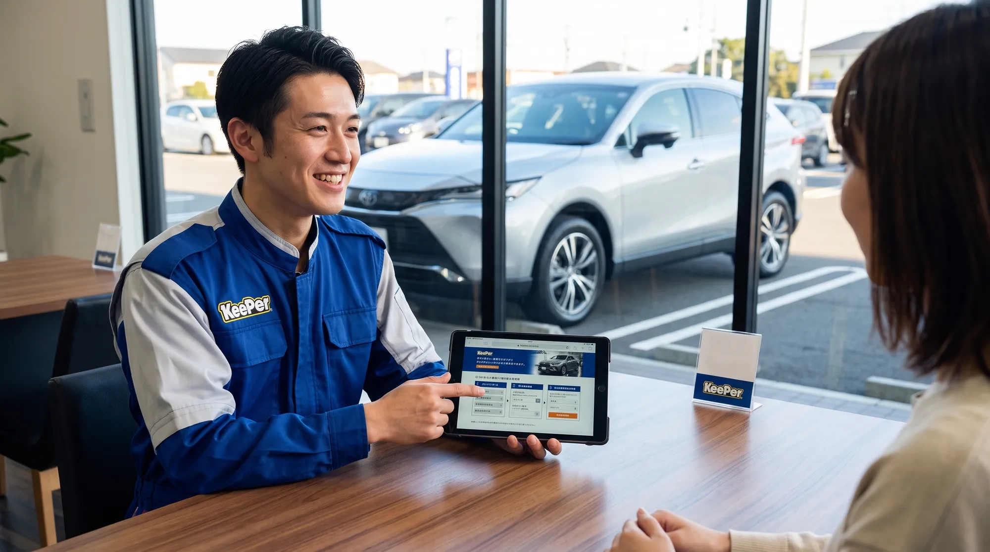 制服姿のスタッフが、デスク越しにタブレット端末を使いながら笑顔でお客様にメニューや予約手順を案内しているシーン。背景の窓の外には施工後のSUVが停まっています。