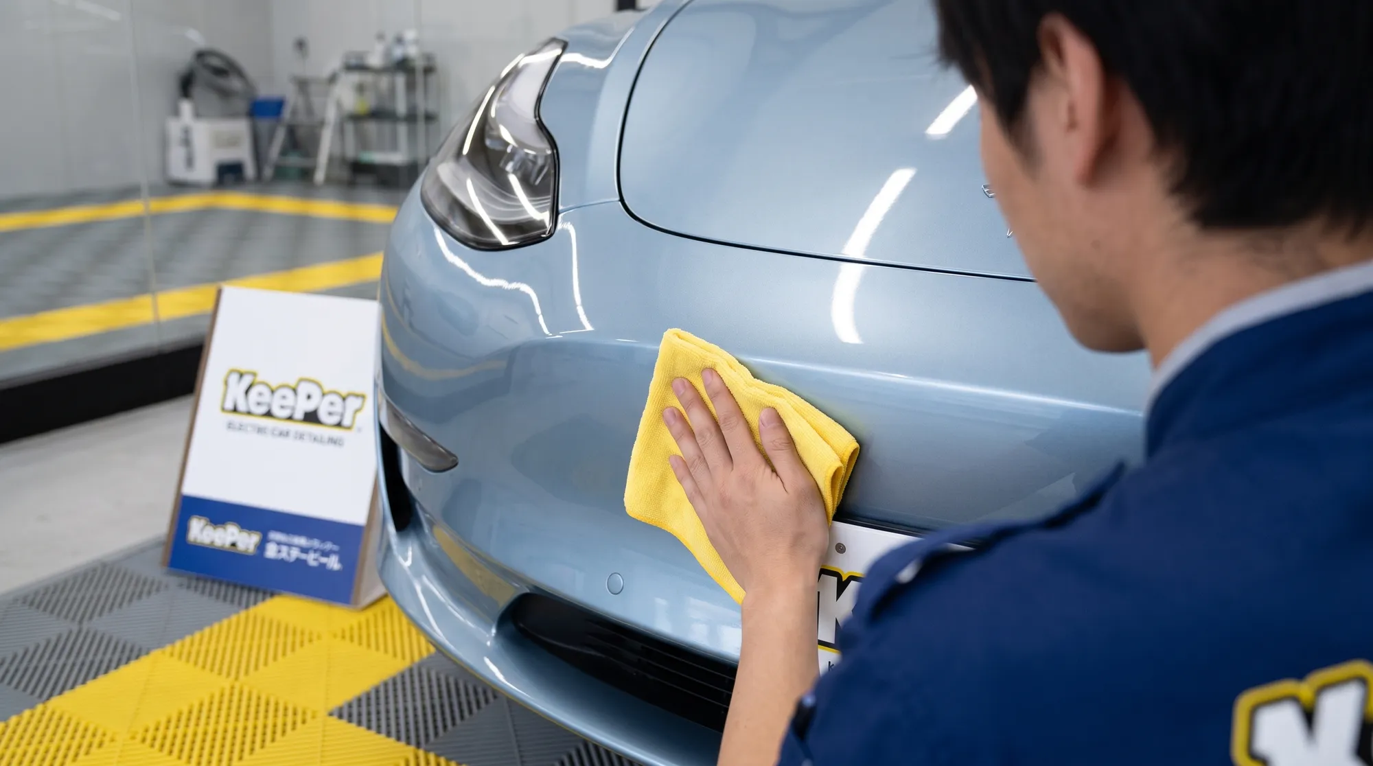 艶やかな淡いメタリックブルーの電気自動車の滑らかなフロントバンパーを、スタッフが黄色のクロスを使って優しく丁寧に拭き上げている手元のクローズアップ。