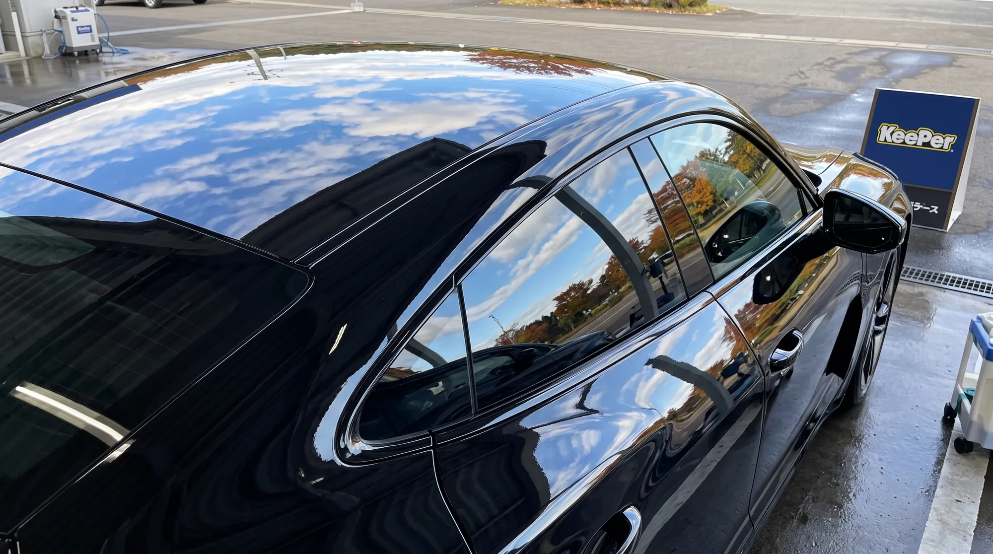コーティング施工直後の漆黒の電気自動車のルーフからサイドミラーにかけてのクローズアップ。鏡のように美しいボディ表面に秋の青空と白い雲が反射している様子。