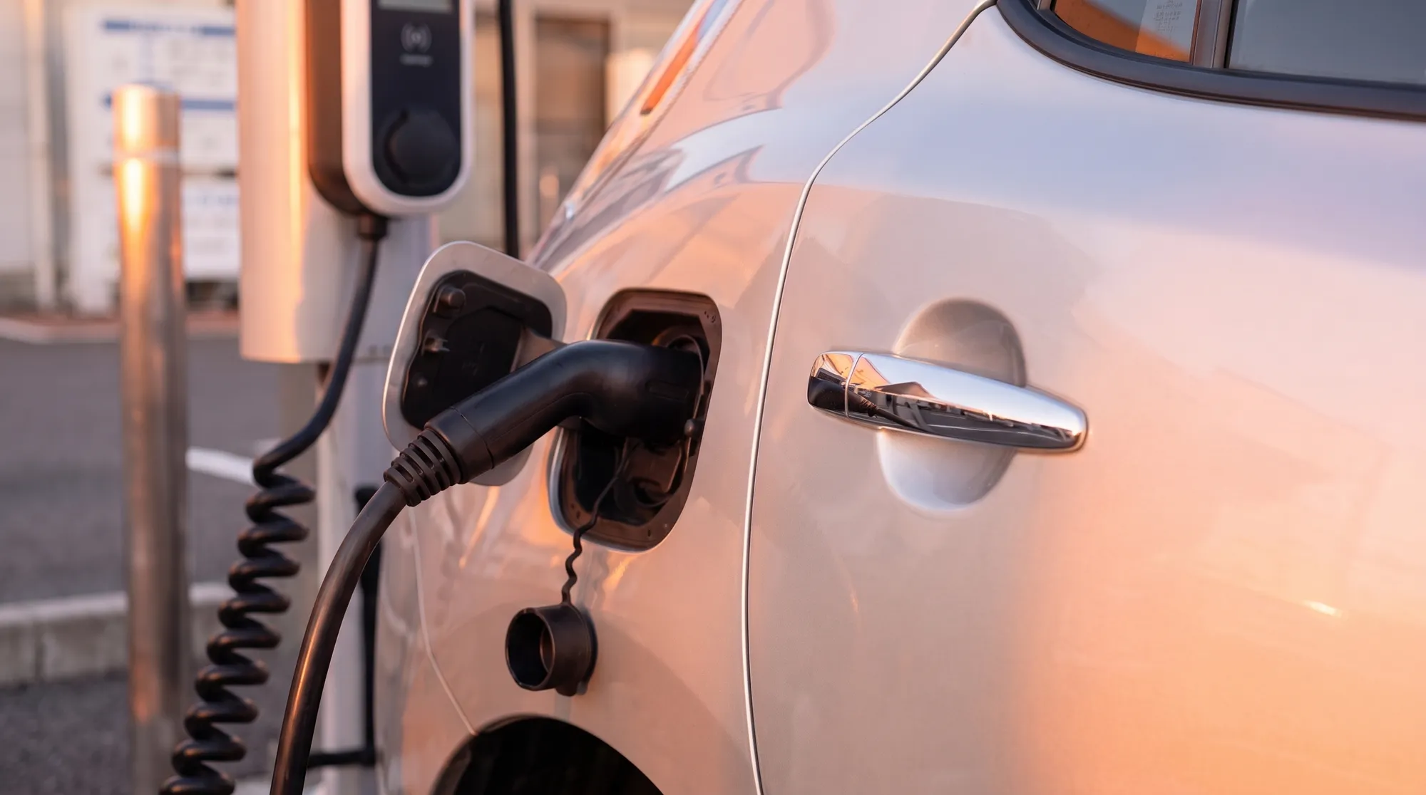 西日が差し込む冬の晴れた午後に、電気自動車の側面に太い充電ケーブルが接続されている充電ポート付近を捉えたクローズアップ。背景には充電スタンドが見える。
