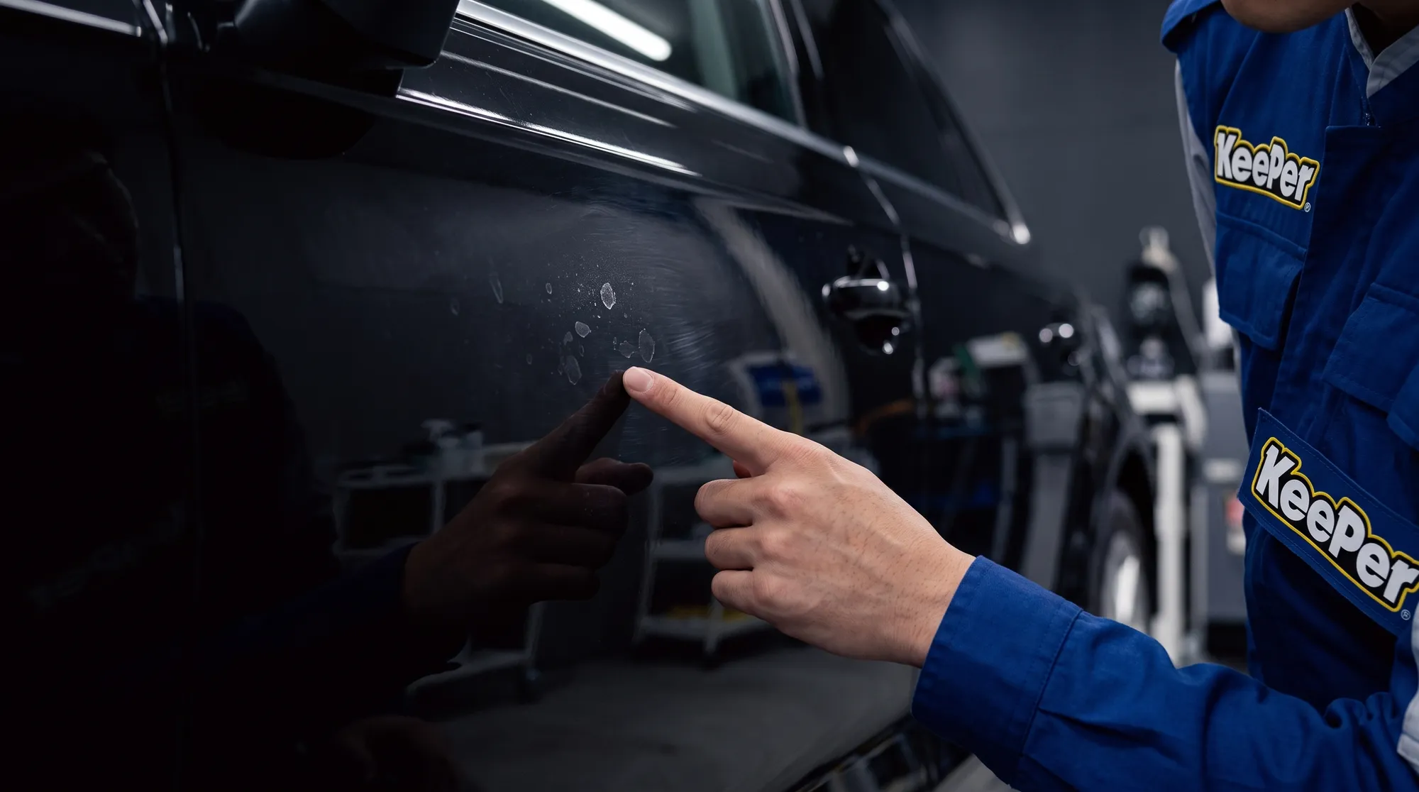 車のドアパネル表面にうっすらと付着した水滴の跡や微細な洗車キズを、作業着姿のスタッフが指差し確認している手元のクローズアップ。屋内施工ブースの照明下での様子。