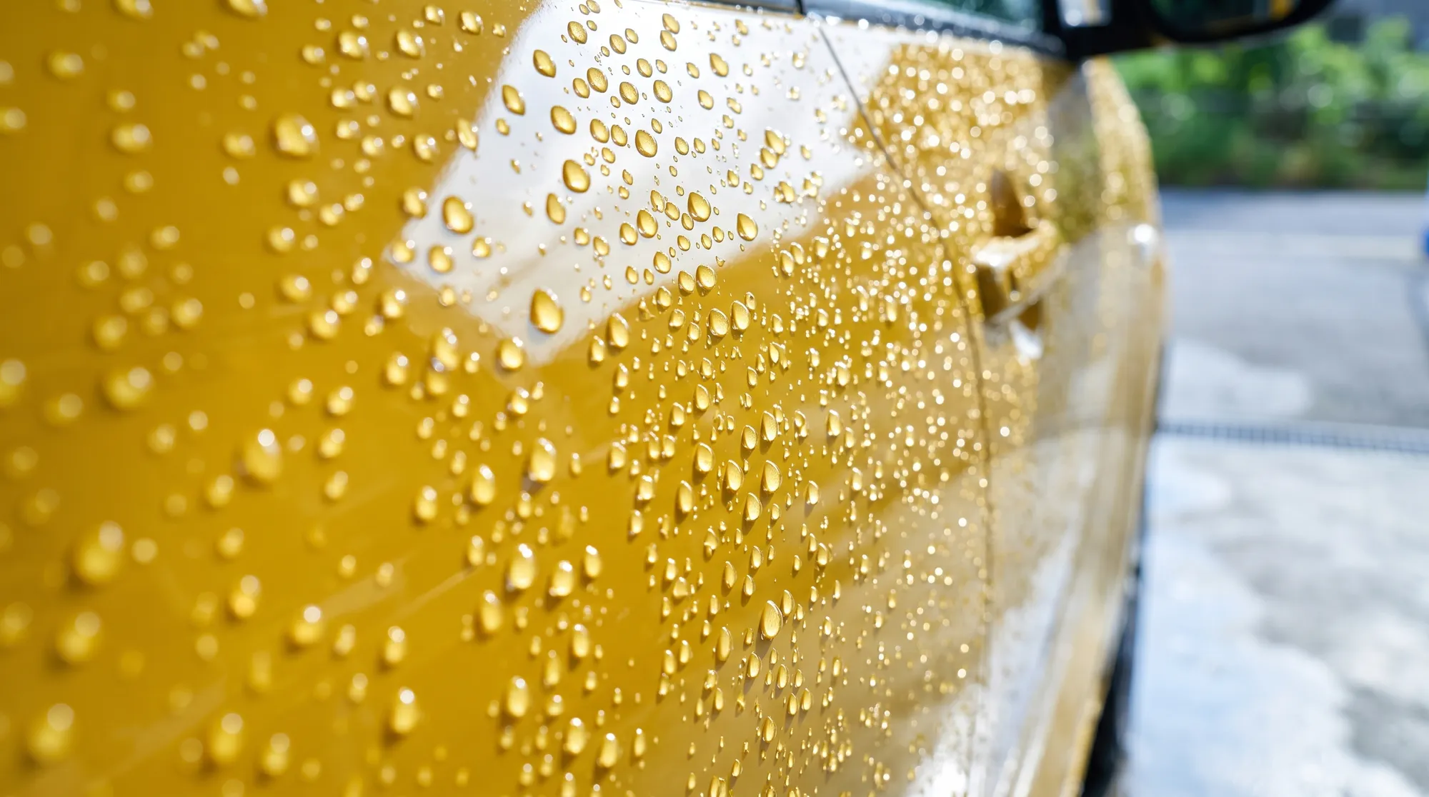 イエローの軽自動車のドアパネルのクローズアップ。コーティング効果で丸まった水滴がボディ上で弾かれ、朝の自然光を反射してキラキラと輝いているみずみずしい様子。