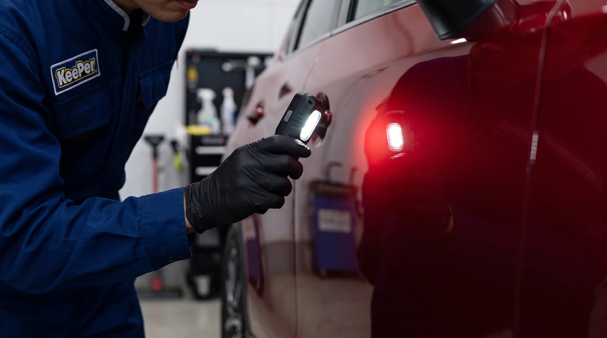 コーティング施工後の車体をライトで照らし、仕上がりを確認するスタッフの手元のクローズアップ。黒い手袋をした手が、艶やかな赤いドアパネルを照らしている様子。