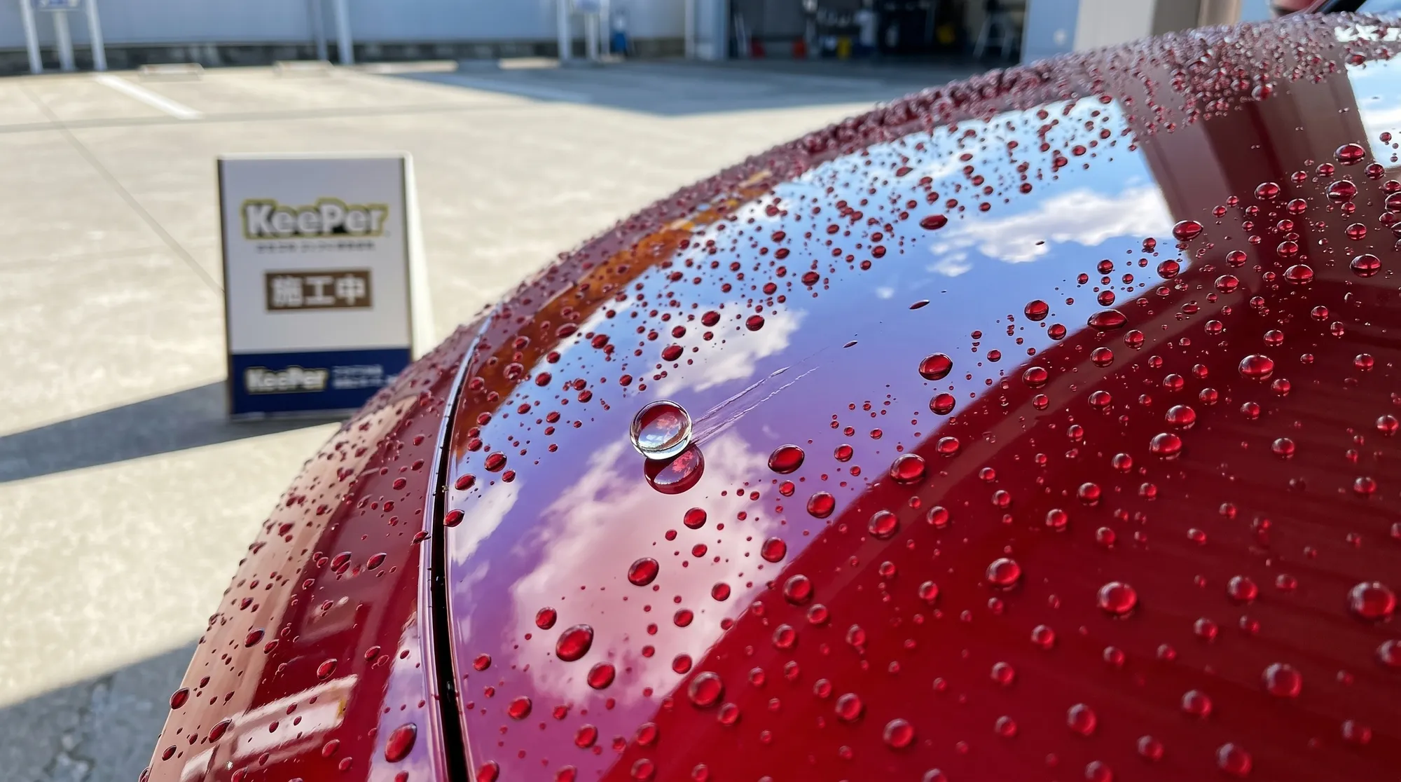 艶やかな赤い車のボディを球状の水滴が滑り落ちる瞬間のクローズアップ。鏡のような表面に青空が映り込み、強力な撥水効果と水弾きの良さがひと目で伝わる様子です。