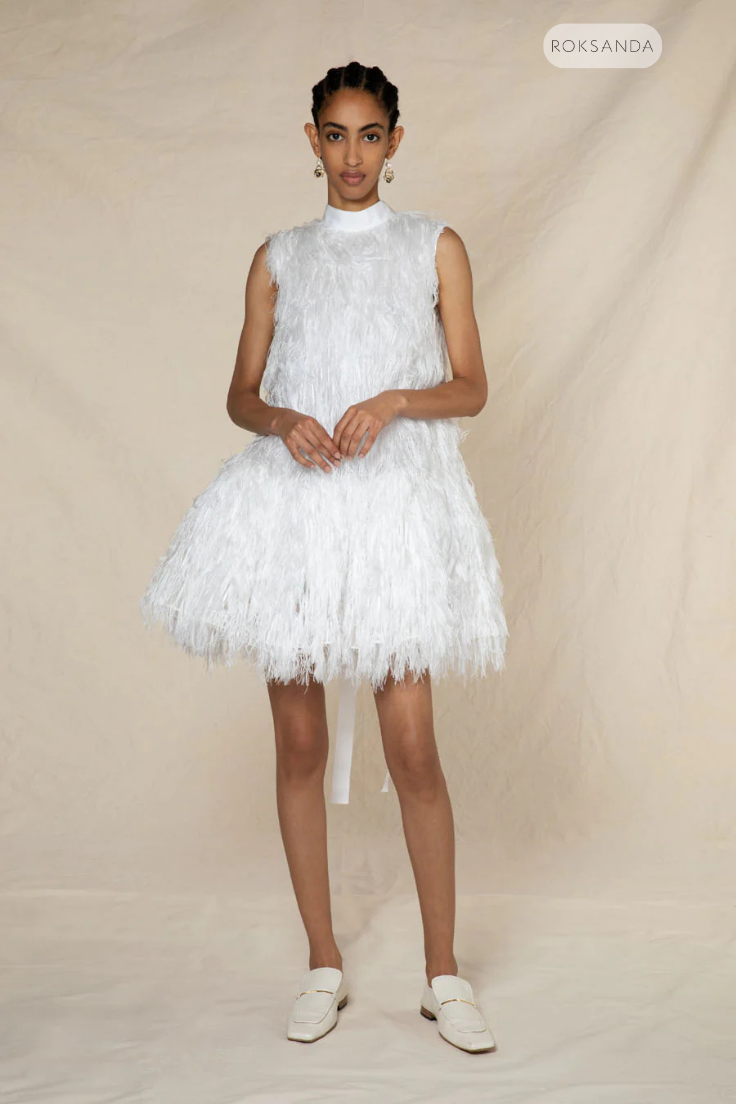 Woman in a white fringed dress stands against a beige backdrop. She wears earrings, white loafers, and exudes a calm demeanor.