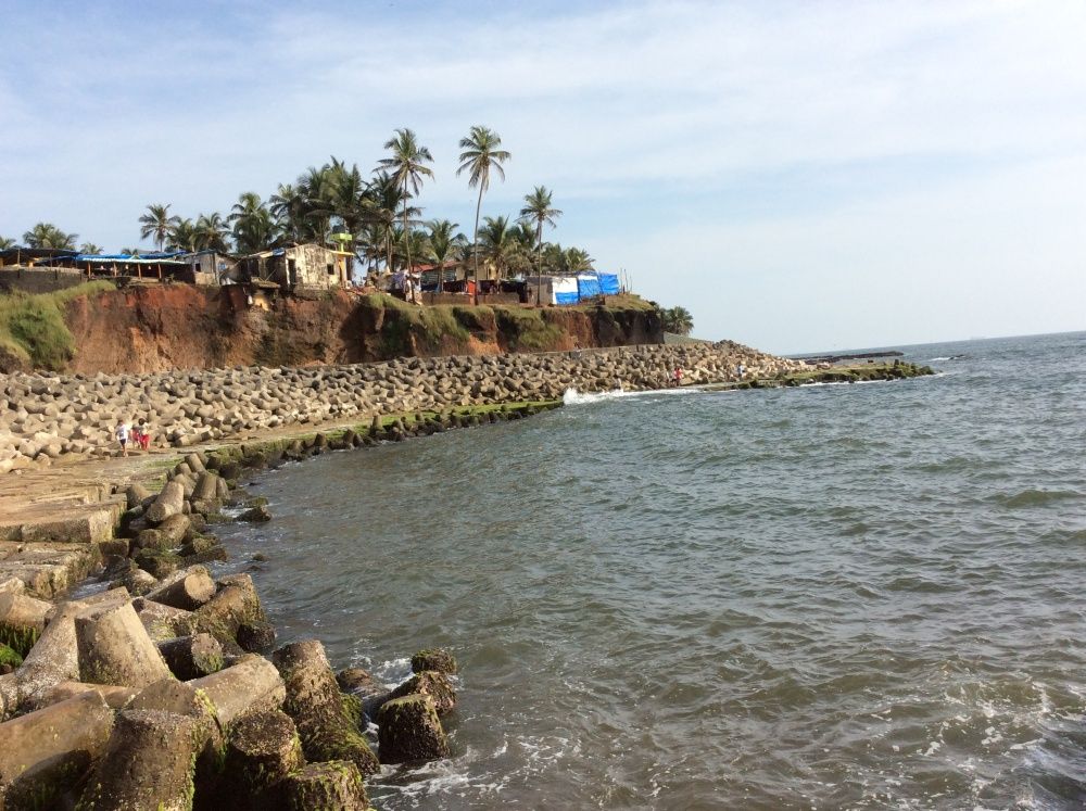 Wide landscape view of Anjuna Beach from above, Goa — showing the full bay, the meeting of red laterite cliffs and white sand, and the expanse of the Arabian Sea stretching to the horizon under a vivid blue sky