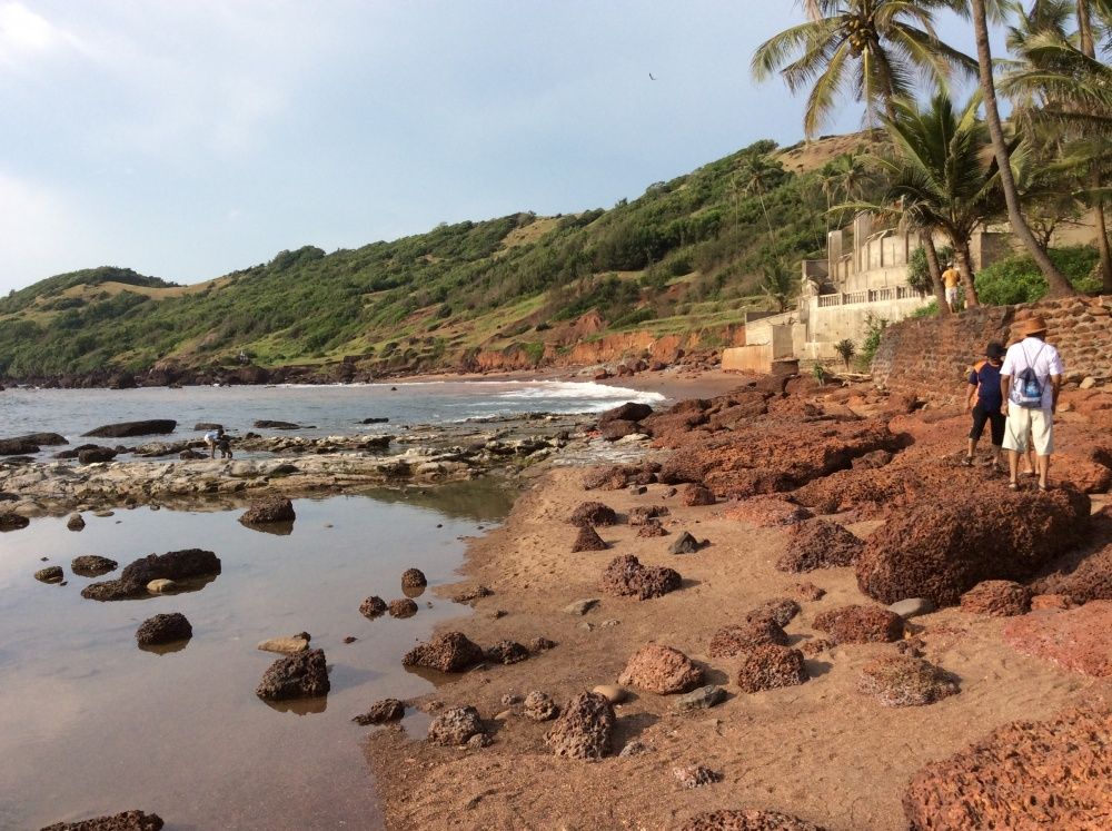 Side view along Anjuna Beach shoreline, Goa — looking along the water's edge where gentle waves wash over the golden sand between red rock outcrops, with palm trees and beach shacks visible along the treeline