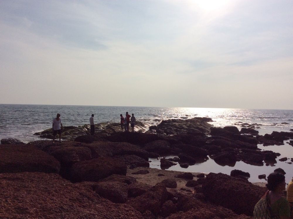 Panoramic view of Anjuna Beach, Goa — the full sweep of the sandy bay with the deep turquoise Arabian Sea, red laterite rocks in the foreground and clear blue sky above