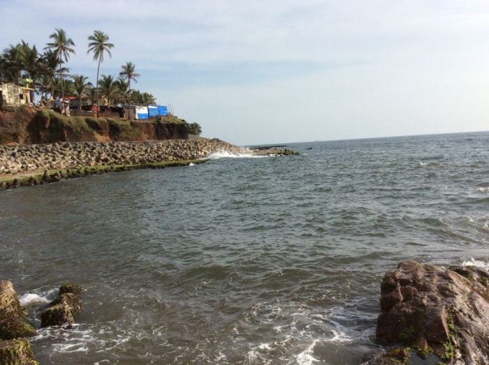 Anjuna Beach view at Goa showing the vivid contrast between the deep red laterite rock formations and the clear turquoise-green water of the Arabian Sea on a bright afternoon