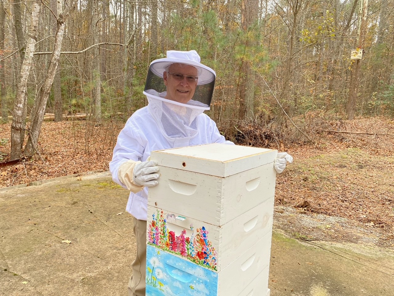 A senior dressed in protection gear poses next to the beehive