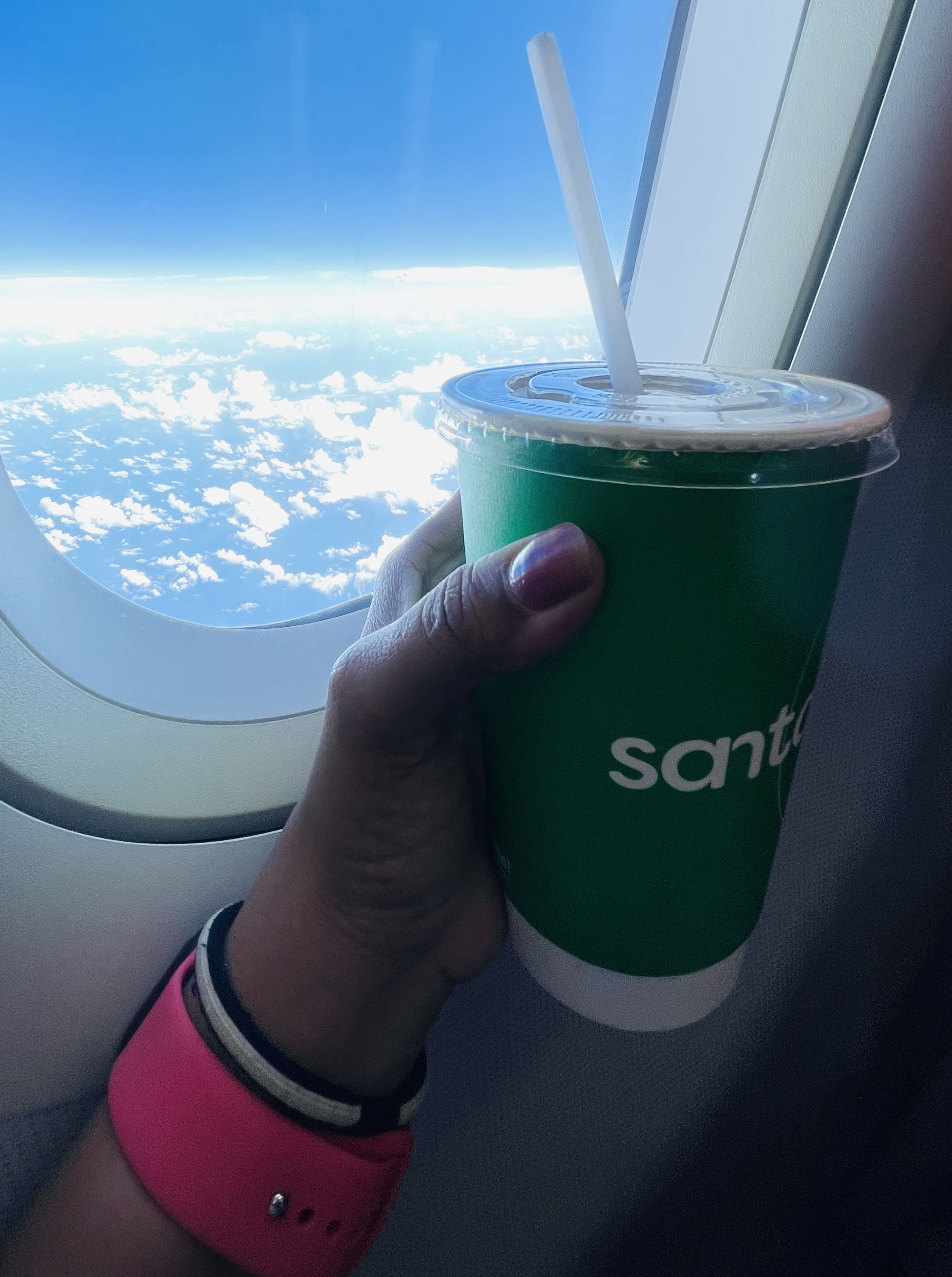 A hand holding a Santan drink cup against the aeroplane window above the clouds