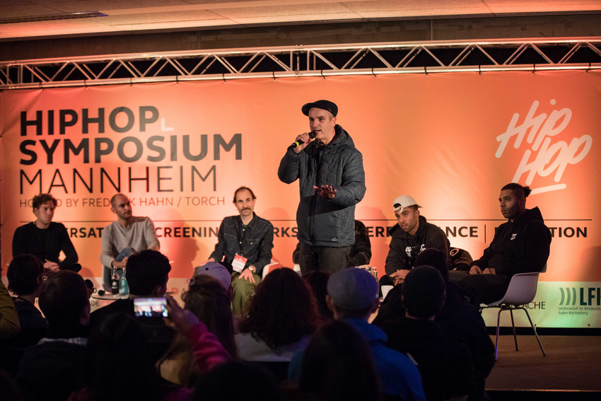 Der Heidelberger Rapper Toni L (stehend) beim Hip Hop-Symposium in Mannheim), links Moderator Jan Possmann vom zeitraumexit, links daneben der Politiker Dr. Danyal Bayaz, ganz rechts Megaloh.