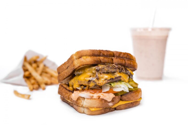 Roman-style burger from M Burger, made with a grilled cheese sandwich for each bun. A portion of fries and a milkshake are displayed in the background.