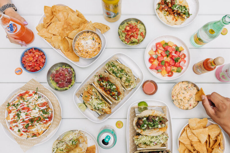 Hands featured enjoying soda beverages and a spread of various Tallboy Taco dishes such tacos, salads, salsas and guacamole