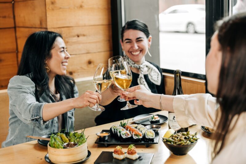 Three women toasting with glasses of sake over sushi, shishito peppers and edamame.