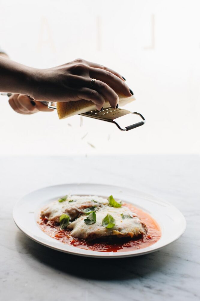 Hand holding a cheese grater and spreading slivers of cheese on a saucy plate of eggplant parmesan from RPM