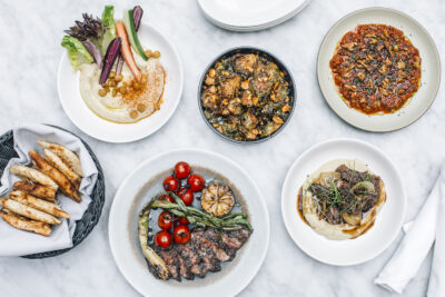 Overhead photo of a variety of dishes including steak, hummus, pita and Brussels sprouts