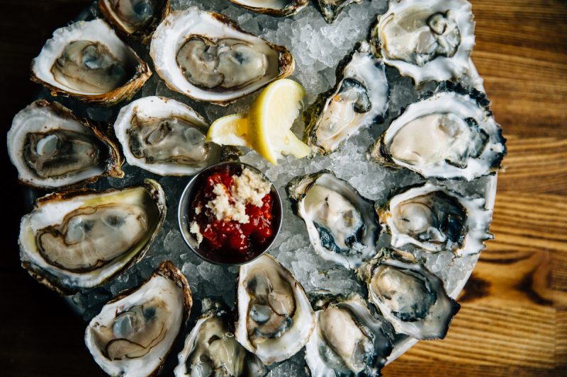 overhead photo of a tray of oysters on crushed ice with cocktail sauce and lemon wedges