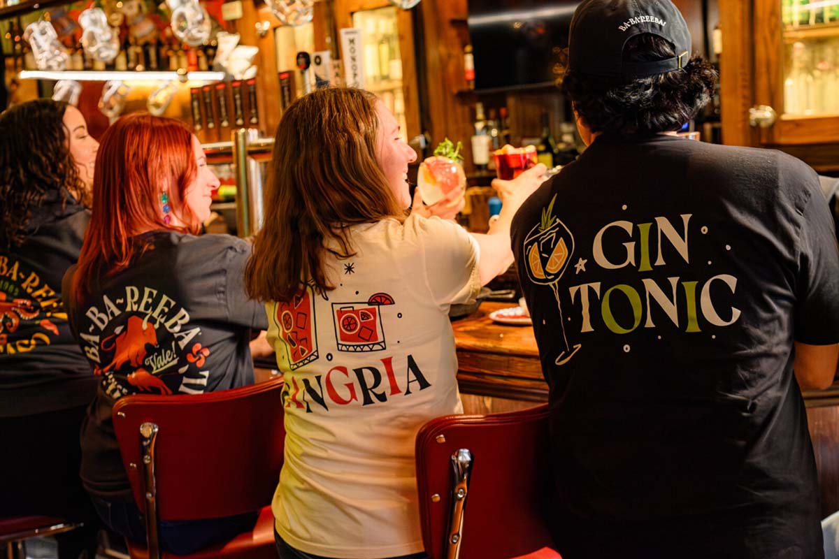 three people sitting on bar stools with their backs to the camera each wearing a cafe baba reeba anniversary shirt