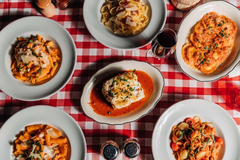Overhead shot of popular pasta dishes and a glass of red wine on a table with red and white checkered table cloth