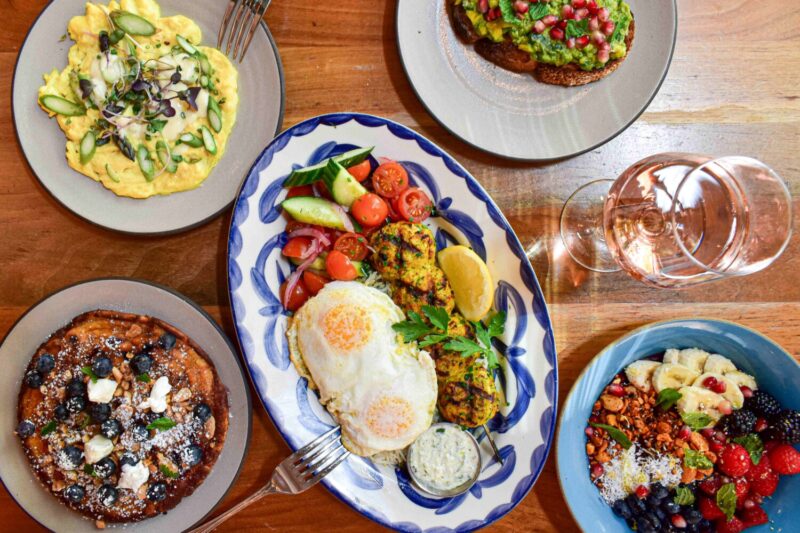 A brunch spread featuring blueberry pancakes, scrambled eggs, fried eggs, chicken skewers, avocado toast, and an acai bowl. 